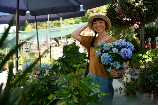 Smiling Asian Woman Shopping at Outdoor Garden Center With Blue Hydrangea Bouquet and Straw Hat