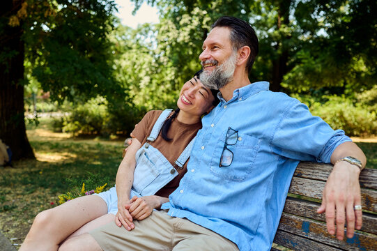 Middle-Aged Man And Asian Woman Relaxing On Park Bench Enjoying Summer Romance And Companionship