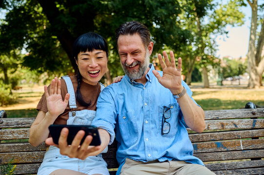Happy Couple Video Calling On Smartphone While Sitting On Park Bench &mdash; Asian Woman And Man Waving