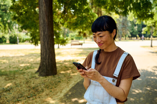 Smiling Asian Woman Using Smartphone in City Park Wearing Overalls on a Sunny Casual Day