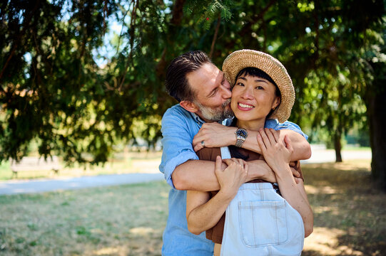Mature Couple Embracing and Kissing in Park: Happy Asian Woman in Straw Hat Smiling