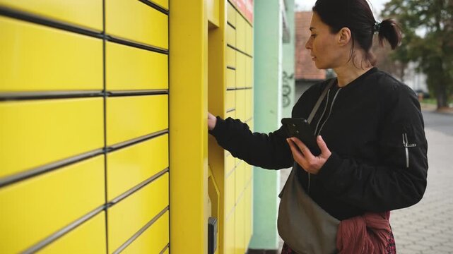 Woman reaching into open compartment of yellow parcel locker, holding smartphone entering parcel code