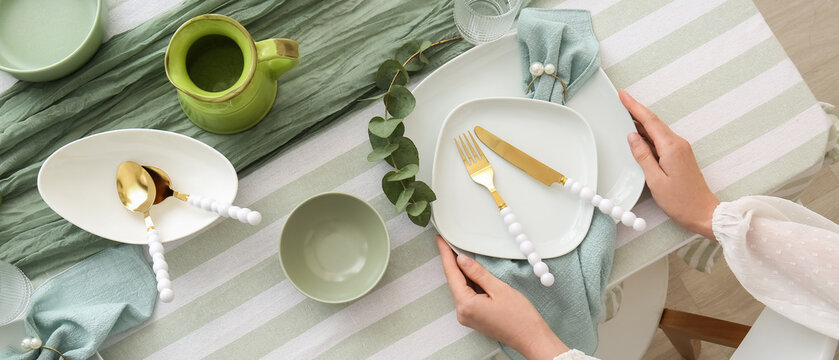 Woman setting a modern dining table with elegant white plates, gold cutlery, and green accents for a special meal