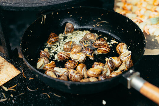 A person prepares snails in a frying pan with garlic and herbs at an outdoor kitchen setting. The cooking process creates steam, and the environment looks vibrant and lively