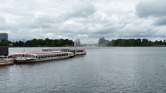 Hamburg, Hamburg, Germany - 22 July 2026: View of the Alster lake in Hamburg with passenger boats moored at the pier and a fountain spraying water in the distance under a cloudy sky.