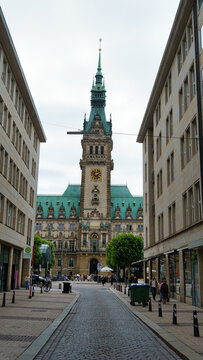 Hamburg, Germany - 22 July 2026: View of the historic Hamburg City Hall with its ornate clock tower and green copper roof, framed by buildings along a cobblestone street.