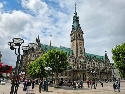 Hamburg, Germany - 22 July 2026: View of Hamburg City Hall with its ornate facade, tall clock tower, and green copper roof, set against a cloudy sky with people walking in the square.