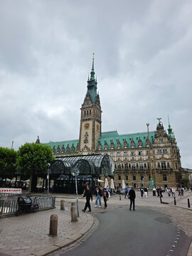 Hamburg, Germany - 22 July 2026: View of Hamburg City Hall with its ornate facade and clock tower, surrounded by a public square with people walking under a cloudy sky.