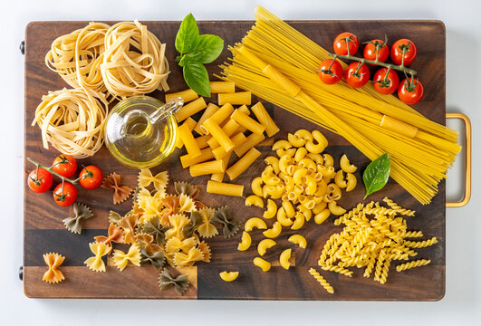 Assorted Italian pasta varieties arranged on a rustic wooden cutting board, including spaghetti, penne, macaroni, farfalle, tagliatelle, fresh basil, cherry tomatoes, red onion, olive oil, 
