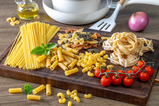 Assorted Italian pasta varieties arranged on a rustic wooden cutting board, including spaghetti, penne, macaroni, farfalle, tagliatelle, fresh basil, cherry tomatoes, red onion, olive oil, 
