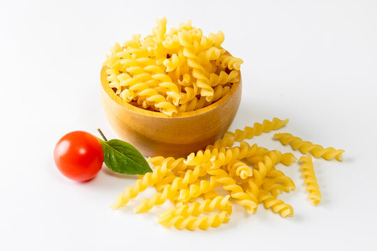 A simple food composition featuring uncooked yellow fusilli pasta in a wooden bowl, surrounded by loose pasta pieces, a fresh tomato, and basil leaf on a clean white background. 