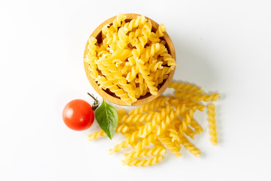 A simple food composition featuring uncooked yellow fusilli pasta in a wooden bowl, surrounded by loose pasta pieces, a fresh tomato, and basil leaf on a clean white background. 
