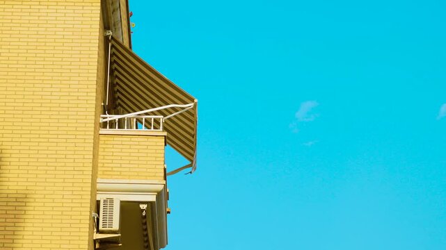 Striped retractable awning on apartment balcony under bright blue sky. Residential building facade with outdoor sunshade and air conditioner. Mediterranean urban lifestyle in Spain