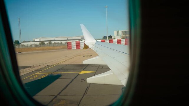 Passenger view through airplane window during pushback from airport terminal. Aircraft wing beside taxiway markings and airport infrastructure. Commercial aviation travel before departure
