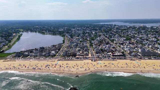 Aerial establishing shot of crowded Belmar beach, ocean, lake Como and coastal town on a sunny day