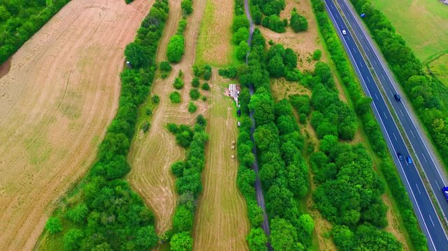 Aerial top-down view of a German autobahn running past a scrubby meadow with hay bales, a stacked timber pile, hedgerows and a golden grain field.