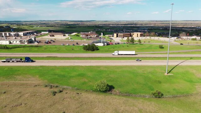Side profile aerial view of an eighteen wheeler heavy rig hauling a loaded flatbed for cargo transport, driving along a scenic midwestern plain with background architecture.