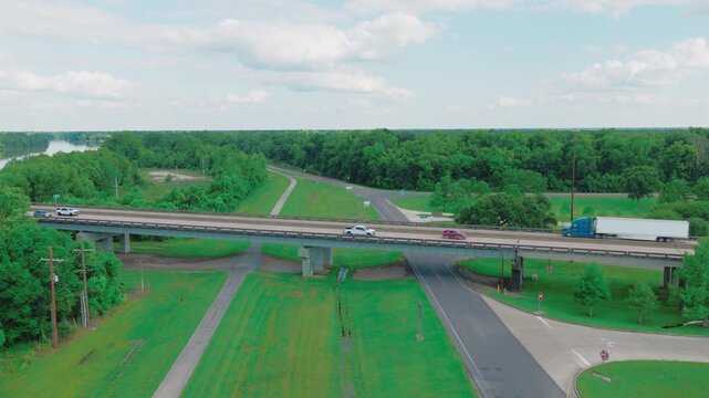 Blue and green commercial semi trucks hauling white trailers on the Interstate 10 elevated bridge over the lush Louisiana swamp.