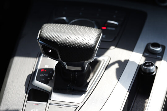 Modern car interior detail shot, automatic transmission gear shift lever displaying "P" for park, console controls, brushed metal finish, bright sunlight creating strong contrasts