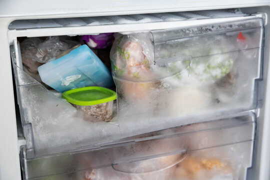 Broken plastic freezer drawer filled with frosted frozen food items and ice, showing damage and heavy ice accumulation inside an old refrigerator freezer compartment, requiring maintenance