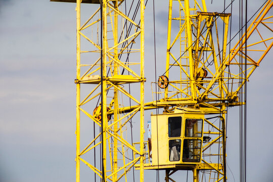Close-up of a yellow construction crane operator cabin and lattice boom structure against a grey sky, indicating ongoing building work at a site for panel house construction.
