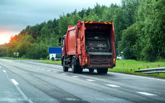 Red garbage truck drives on wet asphalt road through green forest area under overcast sky during day, showing rear compactor mechanism and refuse. Copy space for text