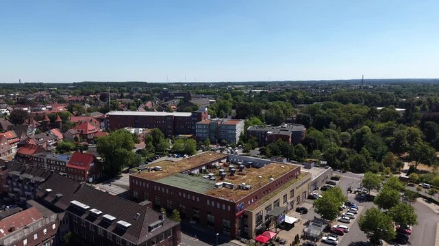 Brick building with green rooftop terrace anchors town center in Dulmen, Germany, red-roofed old buildings and paved parking area line quiet tree-shaded street below. Aerial drone flyover shot