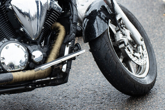 Close-up of motorcycle front section displaying chrome engine components, wrapped exhaust pipe, black wet fender, disc brake on front wheel, parked on damp asphalt road