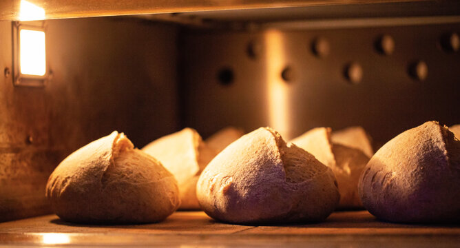Warm loaves artisan bread baking inside industrial oven, golden light illuminating crust bread, traditional baked goods production