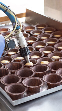 Industrial robotic arm dispensing cupcake batter into paper molds on metal baking tray, automated dessert production line, bakery manufacturing process vertical photo