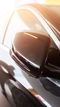 Close up view of side mirror of new dark color automobile parked indoors, reflecting bright sunlight on car body surface, showcasing modern automotive design detail vertical photo