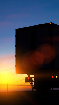 Silhouette of large semi trailer truck driving on road against vibrant sunset sky with bright orange yellow sun light and lens flare, distant horizon, twilight mood vertical photo
