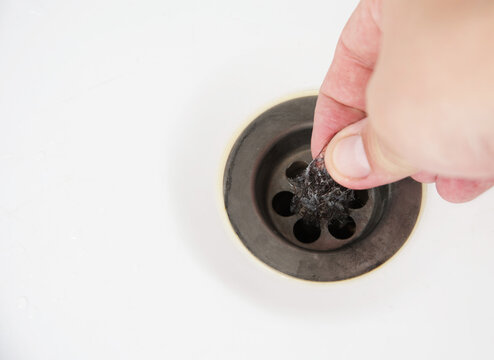 Hand removes clump of dark hair and grime from clogged bathroom sink drain, showcasing common plumbing issue and need for household maintenance and cleaning, copy space