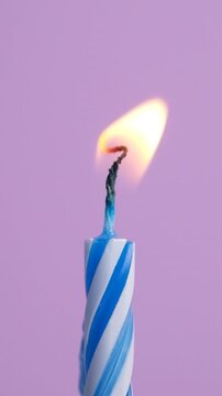Blue and white striped birthday candle burning with a bright flickering flame on a soft purple background. Birthday celebration concept. Slow motion vertical video