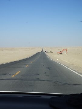 Qinghai, China - 04 August 2026: View of a long asphalt road stretching through the vast desert landscape with an excavator parked on the side under a clear blue sky.