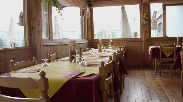 Wide interior shot of a welcoming alpine lodge dining hall in Trentino, Italy. Long wooden tables ready for guests, with large windows framing mountain views
