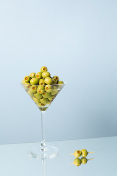 A martini glass holds a large pile of green olives on a blue table
