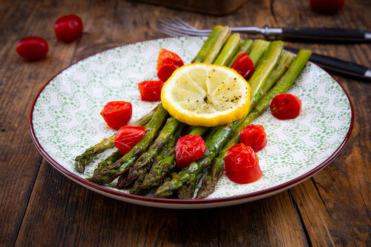 Grilled green asparagus and tomato sit on a dark wooden table