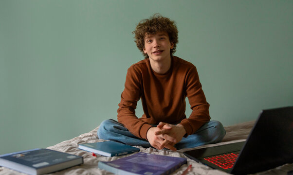Teenage boy sitting on bed studying with laptop and textbooks