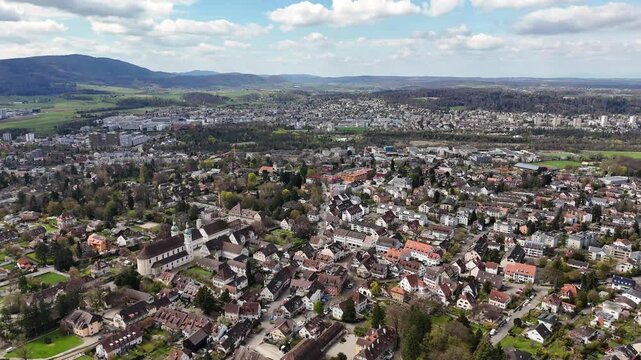 Aerial drone glides over Arlesheim in Switzerland, showing a prominent church, tiled rooftops, residential streets, green fields, and wooded hills beneath scattered clouds.