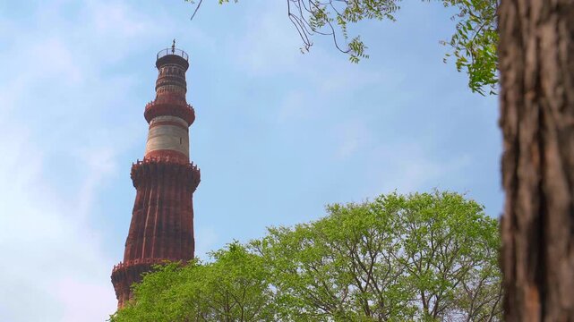 Camera panning left behind a tree trunk to reveal the famous Qutub Minar red sandstone minaret against a blue sky in New Delhi, India.