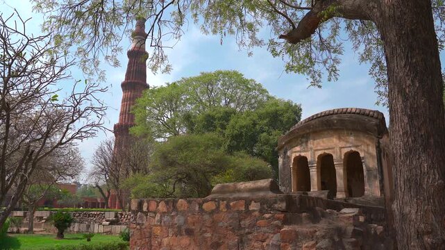 Static wide shot of the iconic Qutub Minar minaret framed by green trees and an ancient stone pavilion at Qutb complex in New Delhi, India.