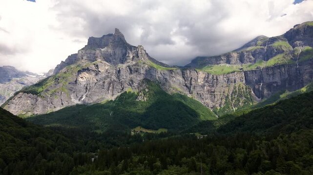 An aerial view moves toward the rugged cliffs of Cirque du Fer a Cheval in France, where thin waterfalls descend into a forested Alpine valley beneath low clouds and shifting sunlight.