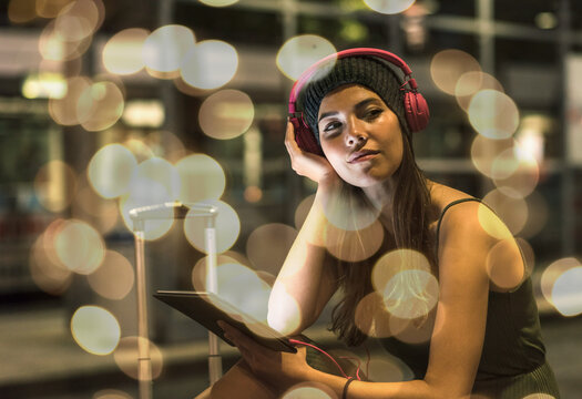 Portrait of young woman with headphones and tablet waiting at station by night