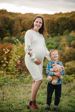 Happy mom and child. Pregnant woman and her little son holding a teddy bear outdoors in autumn. Smiling expecting mother standing with child on a scenic hill.
