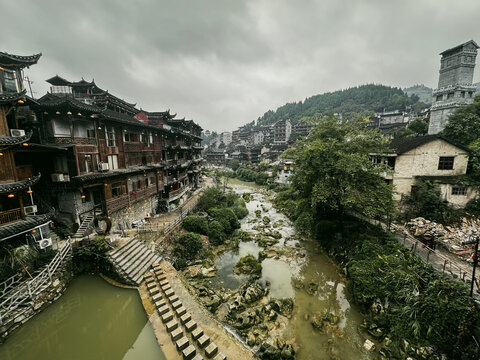 Xiangxi Tujia and Miao Autonomous Prefecture, Hunan, China - 14 October 2024: View of traditional wooden stilt houses along the river in a historic village with lush hills under a cloudy sky.