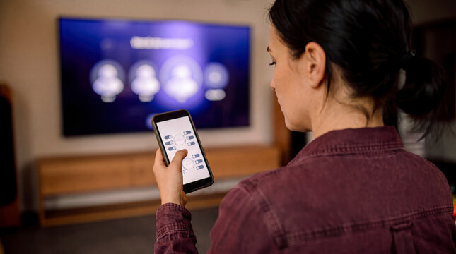 Woman holding smartphone using smart home control application with television in living room
