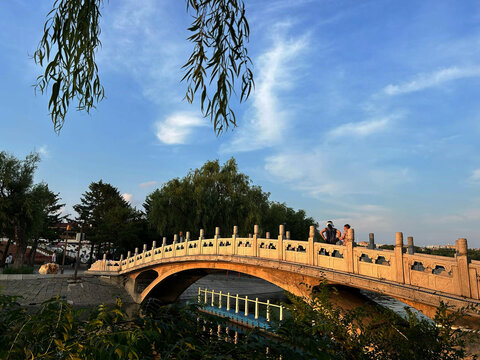 Changchun, China - 03 August 2026: View of Nanhu Park South Lake Park featuring a stone arch bridge with people walking across, surrounded by willow trees under a blue sky.