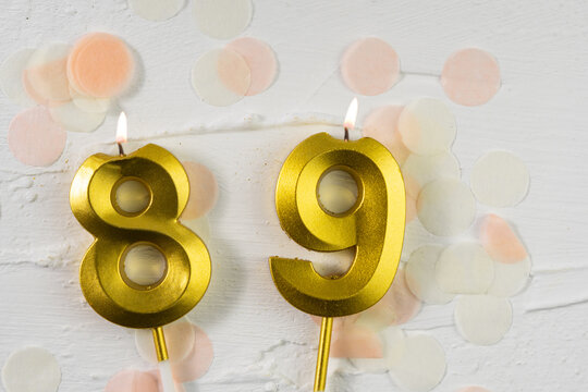Eighty-ninth anniversary celebration. A golden burning candle in the shape of the number eighty-nine on a festive white background with confetti and glitter.