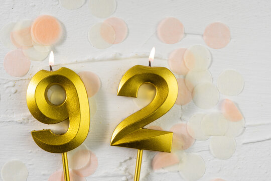 Ninety-second anniversary celebration. A golden burning candle in the shape of the number ninety-two on a festive white background with confetti and glitter.